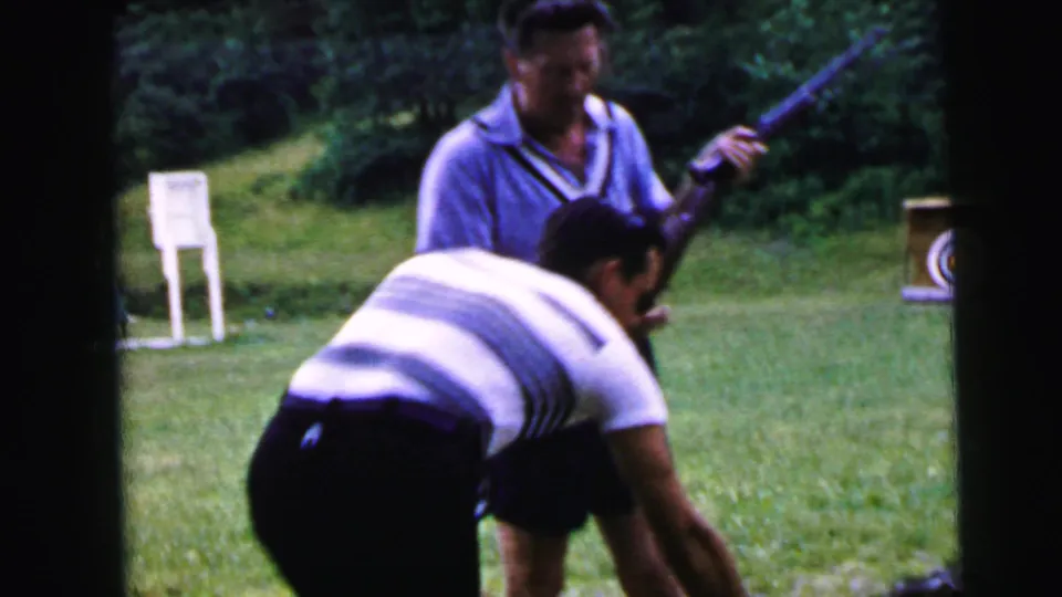At an outdoor shooting range ca. 1964, a man in a striped short-sleeve shirt stoops forward while his companion in a blue polo holds a rifle; a white posted sign and bullseye target stand are visible in the background.