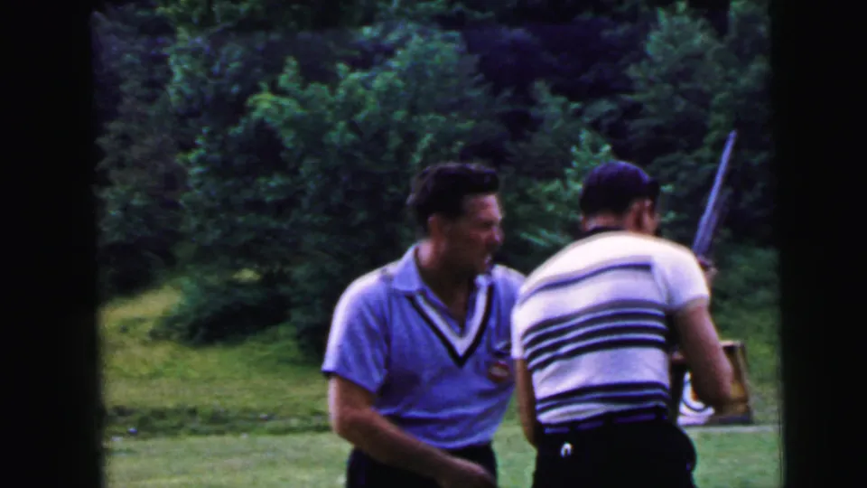 Two adult men together at an outdoor shooting range ca. 1964; the man in the blue polo holds a long rifle while his striped-shirt companion stands at his side; bullseye target stand in background.