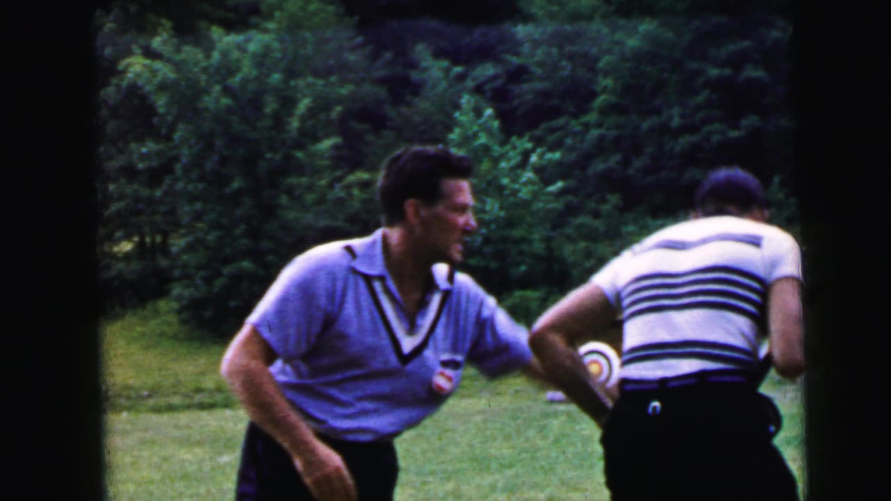 Two men on a grassy field; one in a blue V-neck shirt faces the camera smiling, the other in a striped shirt is seen from behind; circular bullseye target visible at left.