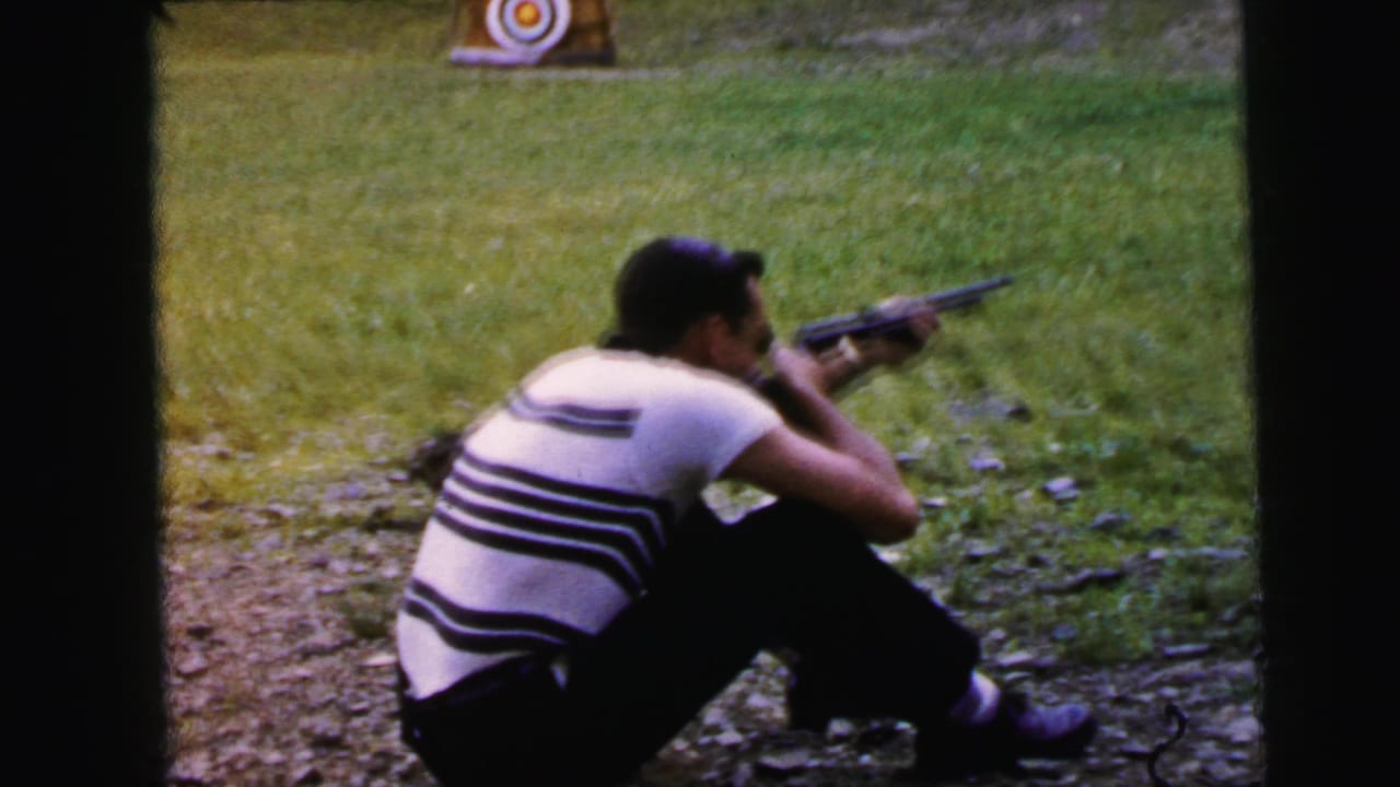 Man in horizontal striped shirt seated on grass aiming a rifle at a bullseye target in an outdoor field, Camden, New Jersey, circa 1964 per metadata.