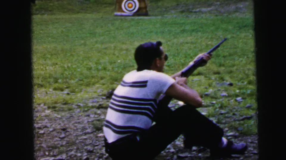 Man in horizontal striped shirt at rest between shots, seated on gravel-edged grass with bullseye target visible at lower right, circa 1964 per metadata.