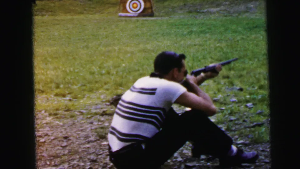 Man in horizontal striped shirt sighting along a rifle at a circular bullseye target in an outdoor field, Camden, New Jersey, circa 1964 per metadata.