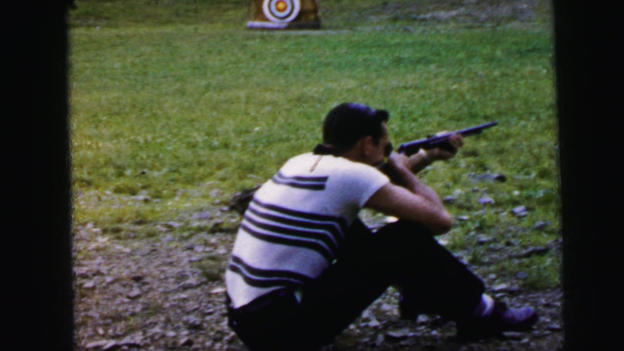 Man in striped shirt seated on dirt, aiming rifle toward a bullseye target visible in the upper right of the frame.