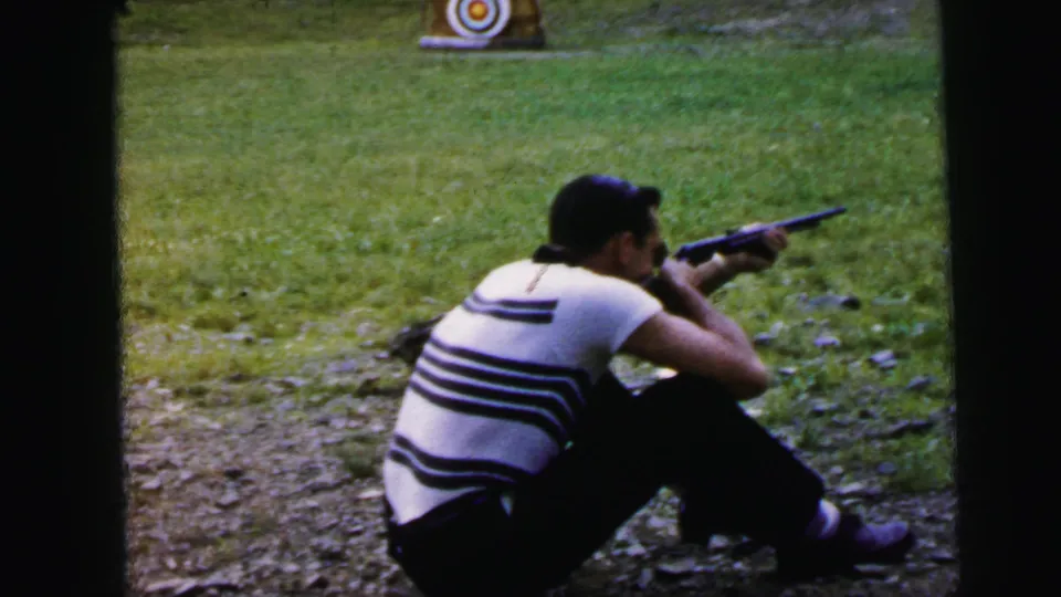 Adult man in horizontal-striped shirt seated at an outdoor shooting area, rifle raised and aimed; circular bullseye target on stand in background; 1964 8mm home movie, Camden, New Jersey.