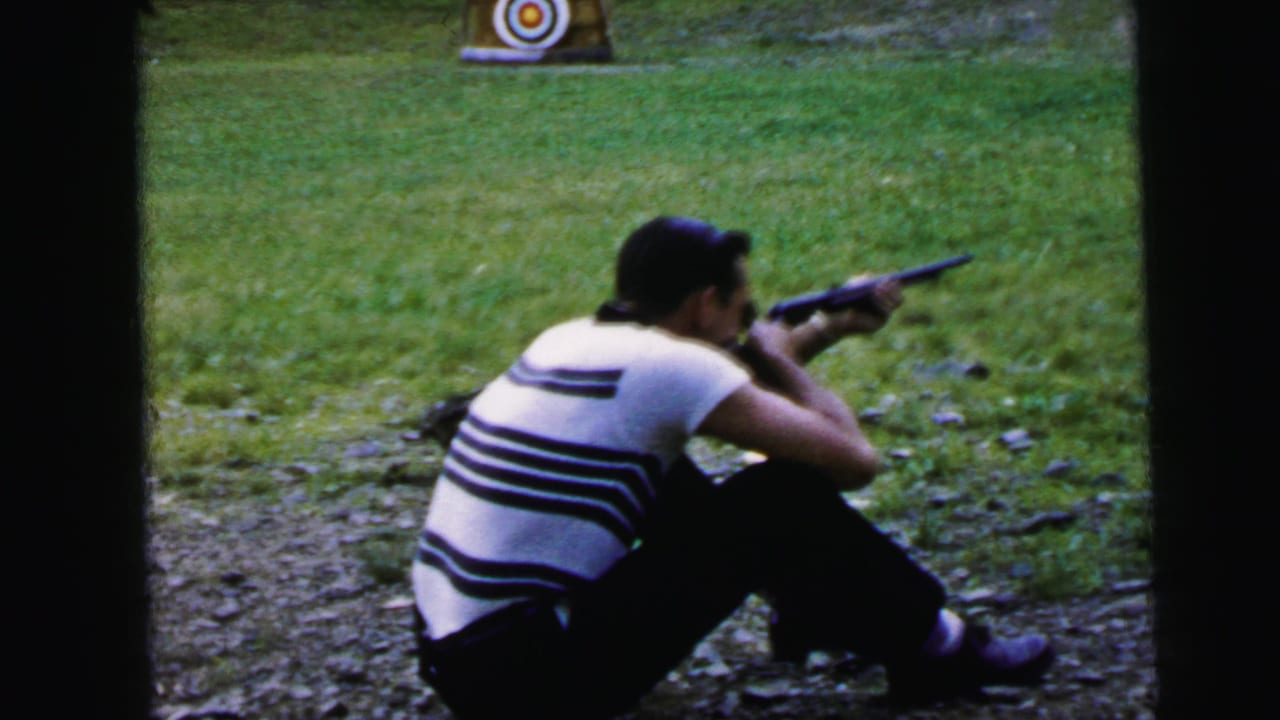 Adult man in black-and-white striped shirt seated and aiming a rifle at a bullseye target; outdoor shooting area with grassy field behind; 8mm home movie, Camden, New Jersey, 1964.