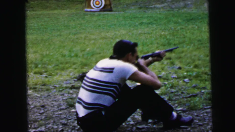 Adult man in black-and-white striped shirt seated and aiming a rifle at a bullseye target; outdoor shooting area with grassy field behind; 8mm home movie, Camden, New Jersey, 1964.