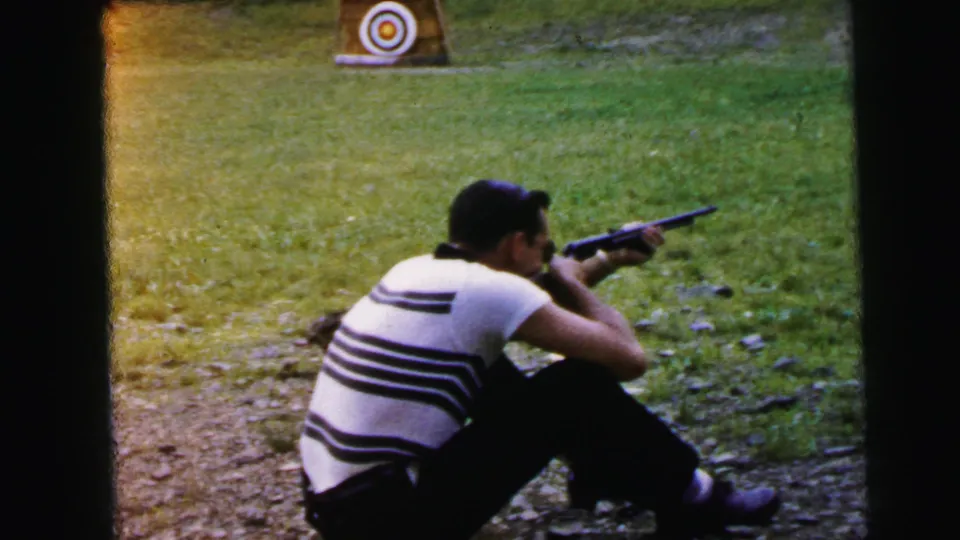 Man aiming rifle outdoors; a second paper bullseye target on stand visible in the foreground left; warm orange dye drift at frame edge is characteristic 8mm film degradation; 1964 footage.