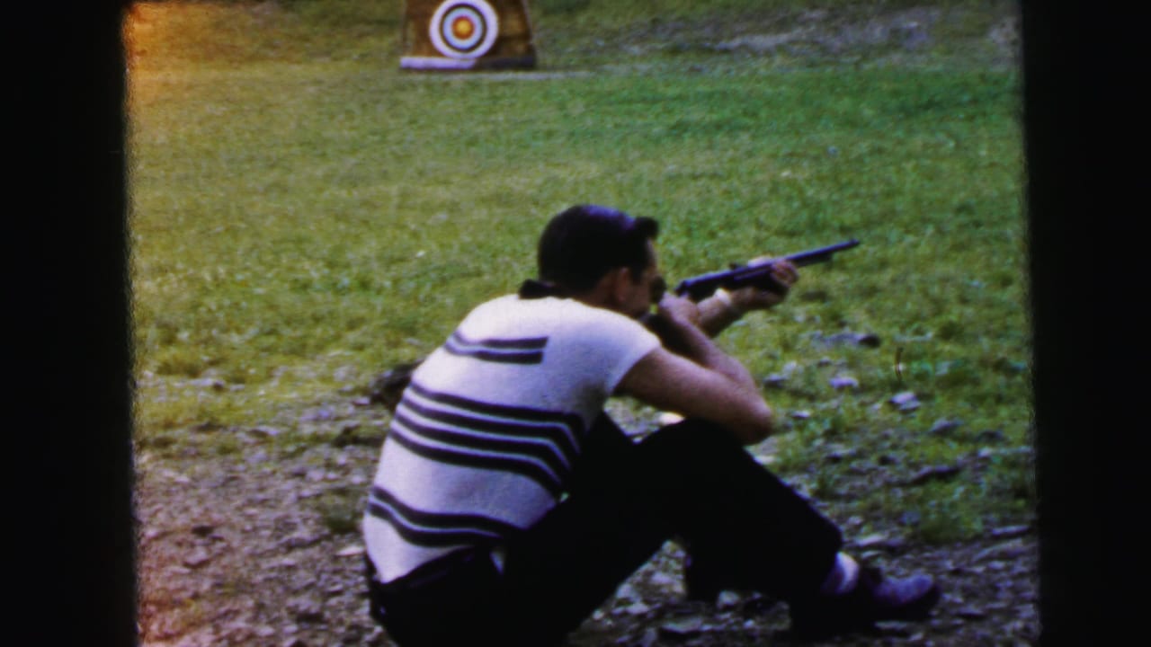 Man in striped shirt seated on gravel, aiming a rifle toward a circular bullseye target on a grass range, seen from behind.