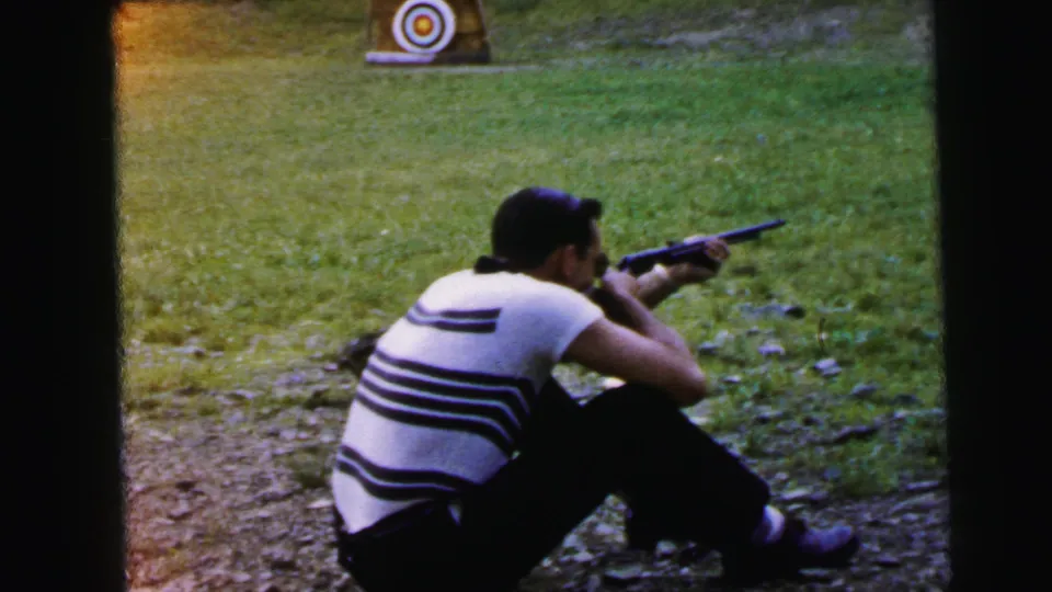 Man seated on gravel at an outdoor shooting range, aiming a rifle toward a circular bullseye target; 8mm home movie, likely 1964, Camden, New Jersey.