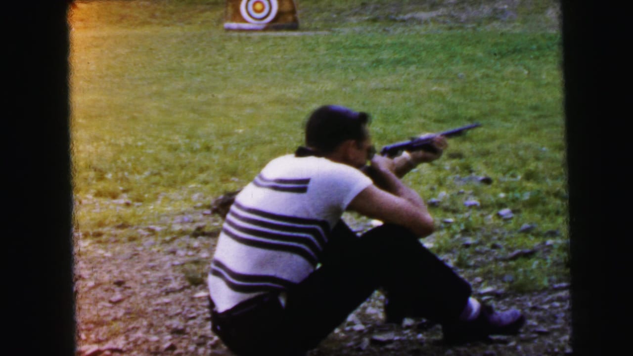Man in striped shirt aiming rifle at a circular bullseye target from a seated position on gravel; 8mm home movie, 1964, Camden, New Jersey.