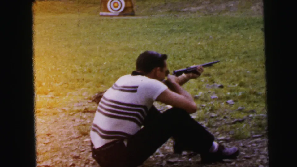 Same shooter seen through strong amber-orange dye-coupler fade; seated aim on gravel at outdoor range, 8mm home movie, 1964.