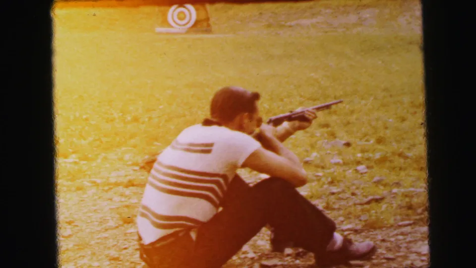 Profile view of man aiming rifle at an outdoor range; continued amber dye-fade typical of unrestored 1960s color reversal stock, 8mm home movie.