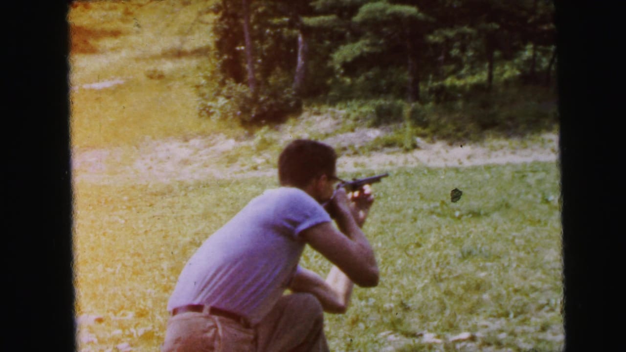 Man crouches in a grassy field aiming a rifle, filmed on 8mm color stock in what is recorded as Camden, New Jersey, 1964; warm color shift visible at frame edge.