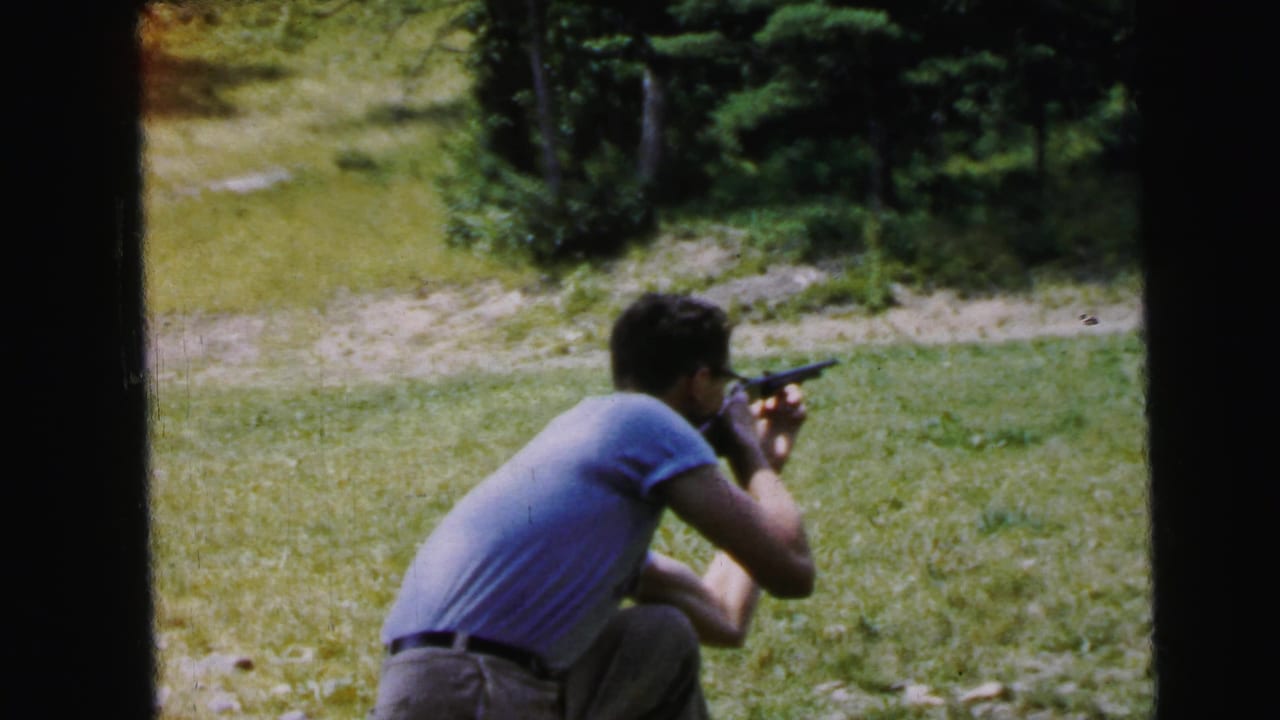 Man crouches aiming a rifle in a grassy field, viewed from behind; 8mm home movie footage recorded as United States, 1964; cooler, more neutral color palette than preceding frame.
