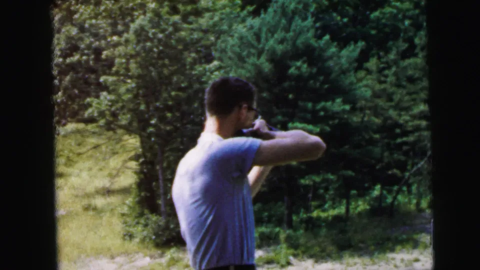Man stands in side profile aiming a rifle, dense conifers behind him; 8mm color home movie with orange exposure flare at frame edge, recorded as Camden, New Jersey, 1964.