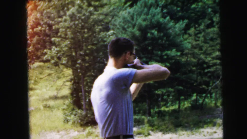 Man stands in profile aiming a rifle in a rural outdoor setting; dense conifers visible behind; faded 8mm home movie color, recorded as United States, 1964.
