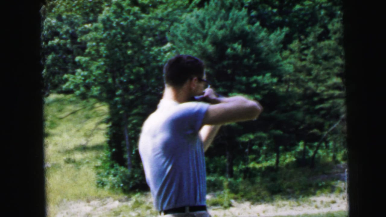 Adult man in glasses and a light shirt aims a rifle outdoors in 1964 Camden, New Jersey home-movie footage.