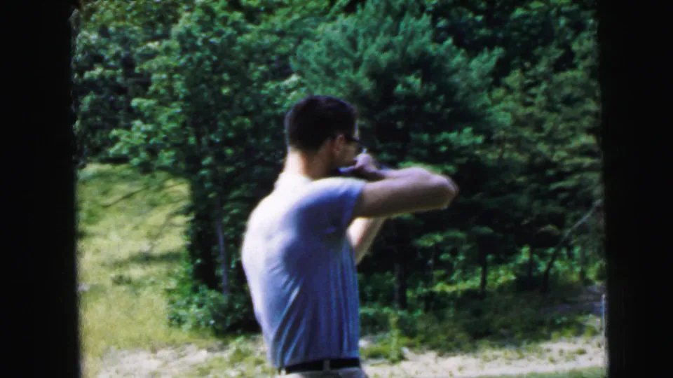 Adult man in glasses and a light shirt aims a rifle outdoors in 1964 Camden, New Jersey home-movie footage.
