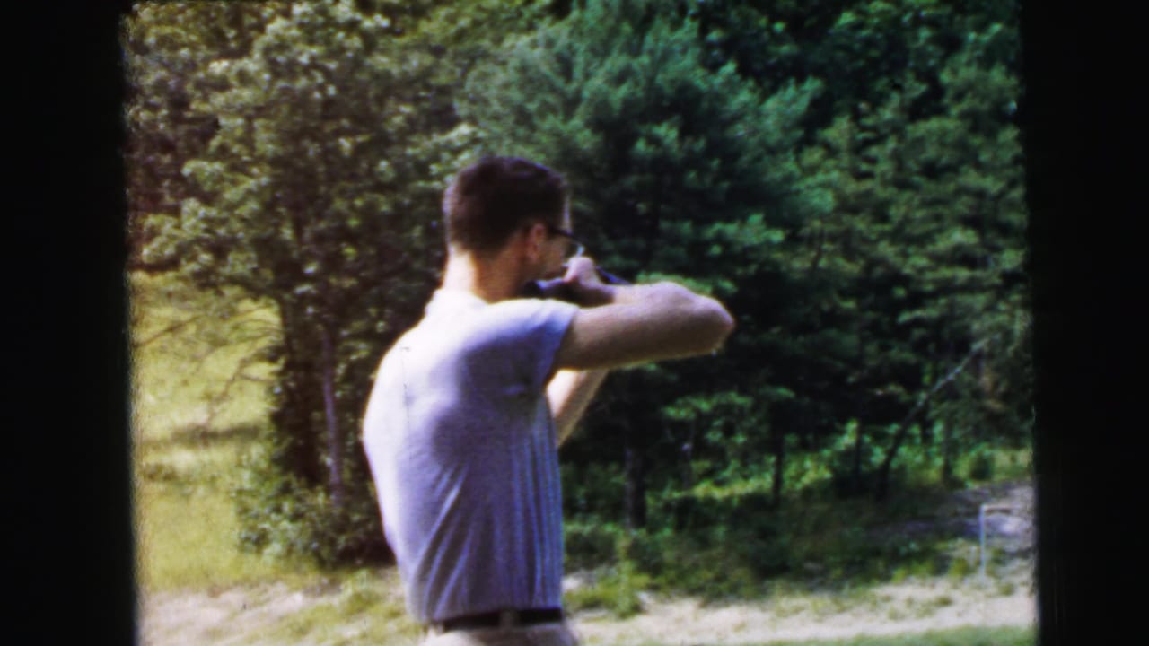 Man holds a rifle to his shoulder beside sunlit trees.