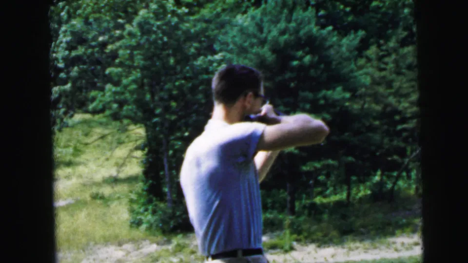 Adult man in a light shirt aims a rifle from a standing position in a wooded clearing in 1964 Camden, New Jersey footage.