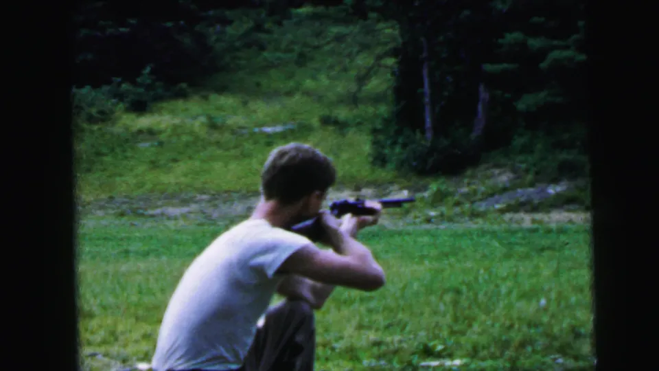 Adult man crouches or kneels as he aims a rifle across grass toward trees in 1964 Camden, New Jersey home-movie footage.