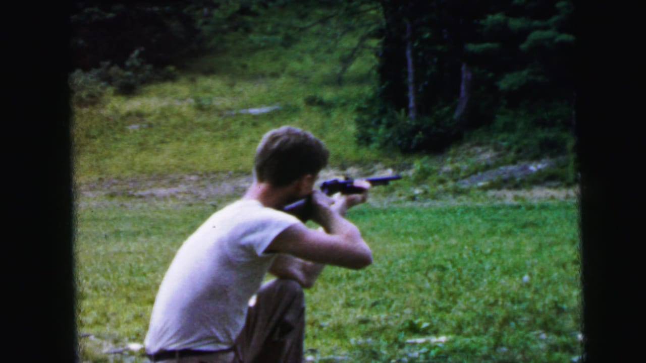 Young man in white t-shirt kneeling on bare earth, aiming a scoped rifle toward the right of frame, green grassy slope and tree line behind.