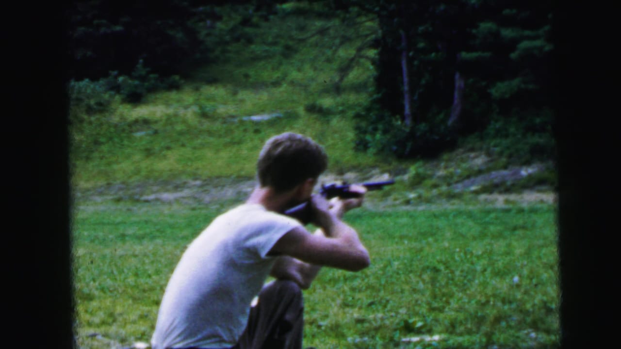 Young man holding a rifle to his shoulder in an outdoor grassy setting, seen from behind; framing and 8mm film stock consistent with home-movie footage, circa 1964.