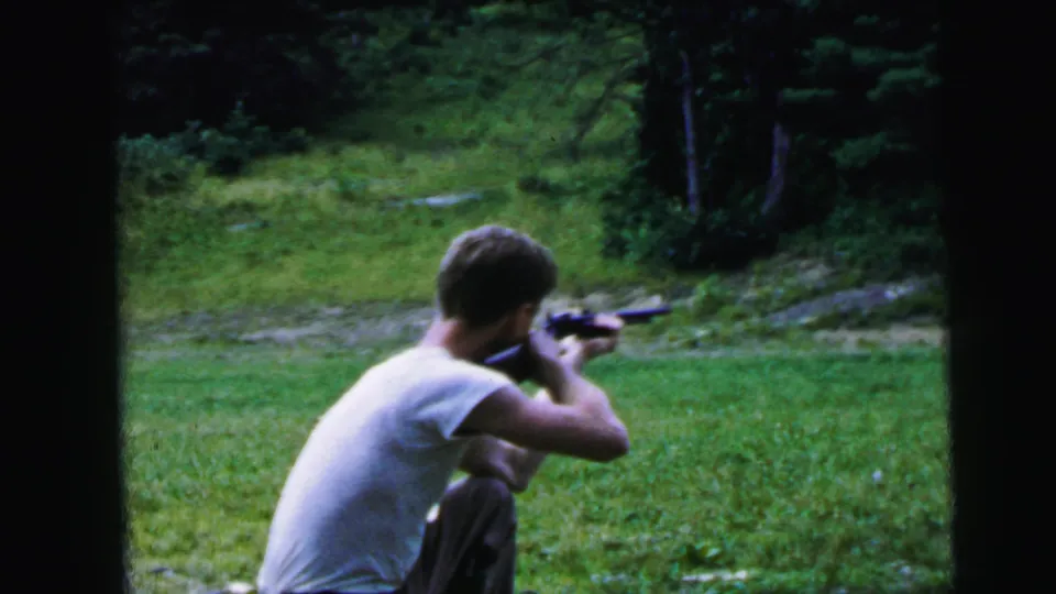 Young man holding a rifle to his shoulder in an outdoor grassy setting, seen from behind; framing and 8mm film stock consistent with home-movie footage, circa 1964.