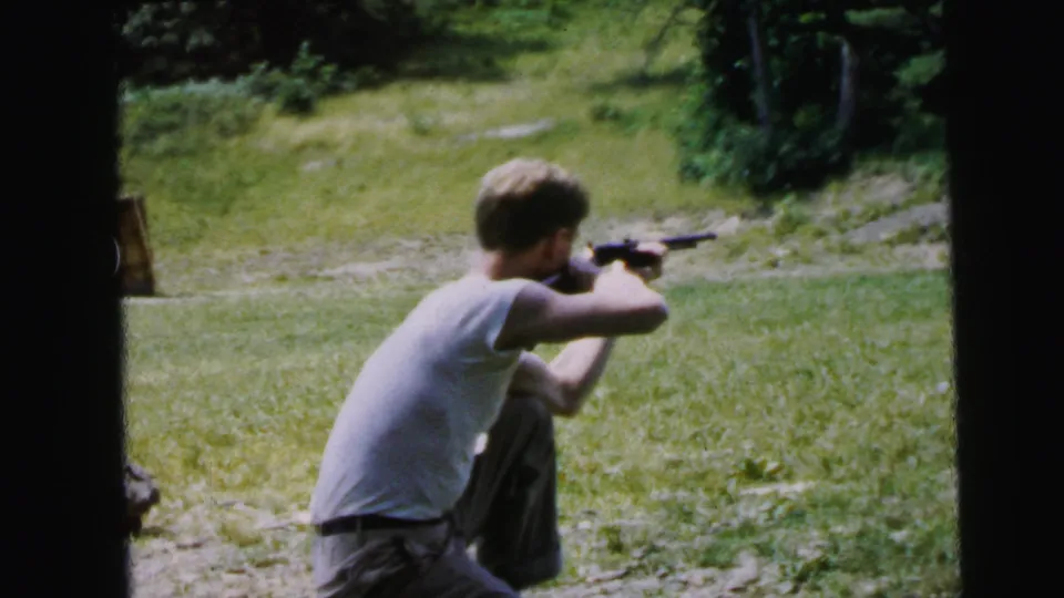 Young man in three-quarter profile kneeling and aiming rifle in grassy outdoor setting; framing reveals more of the subject's right side than earlier frames; circa 1964.