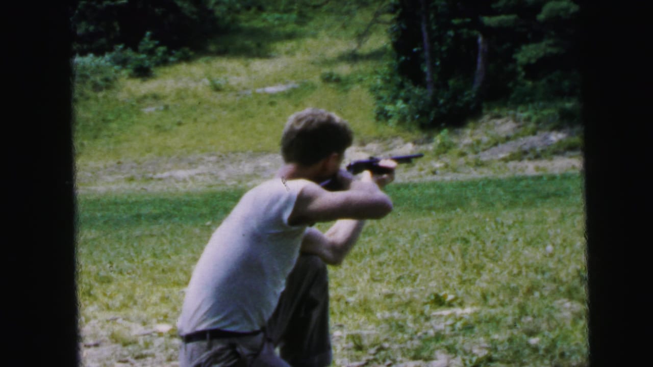 Young man in white t-shirt standing and aiming a rifle across a grassy outdoor field, 8mm frame with dark borders.