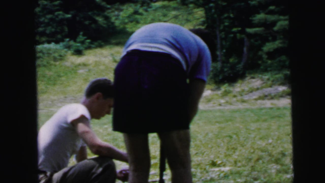 Two figures at an outdoor shooting area: young man in white t-shirt kneeling while adult in blue shirt bends over him.