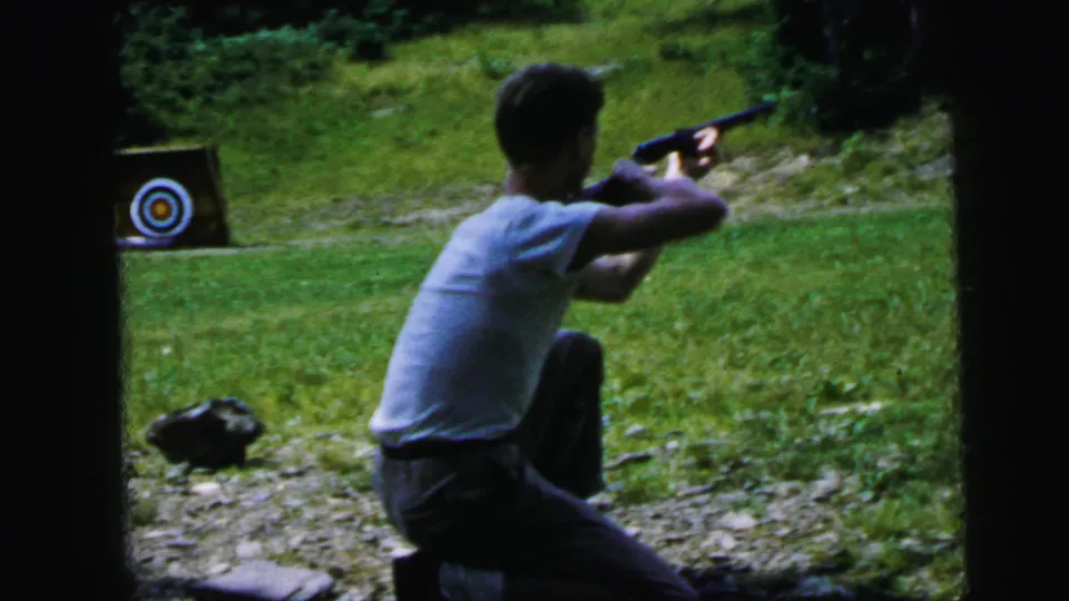 Young man in kneeling stance aiming rifle at a circular bullseye target, outdoor informal range, 8mm home movie, circa 1964.