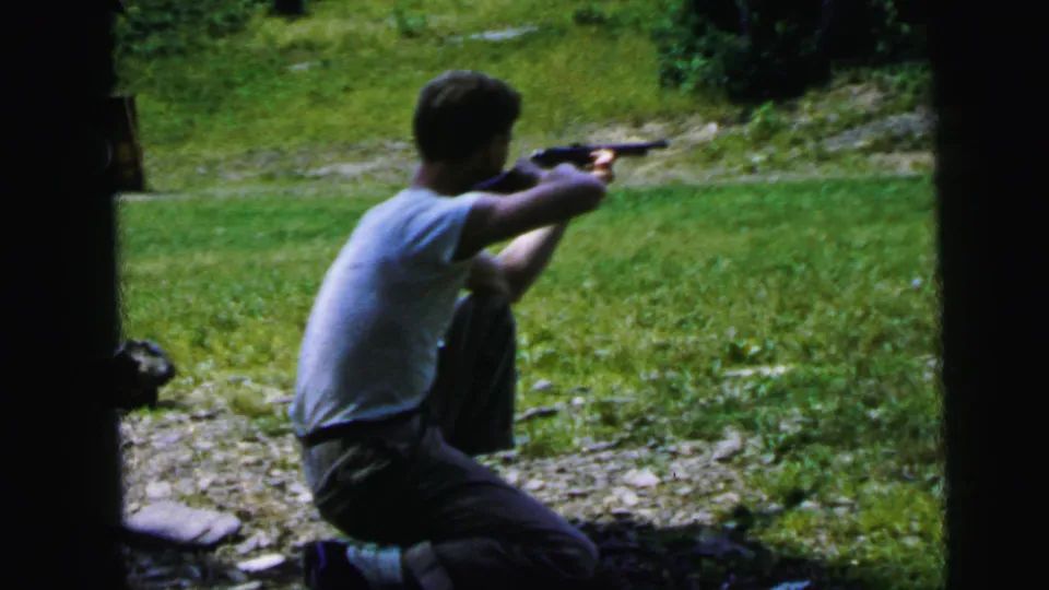 Young man firing or aiming rifle from kneeling position at outdoor range, 8mm home movie, United States, circa 1964.