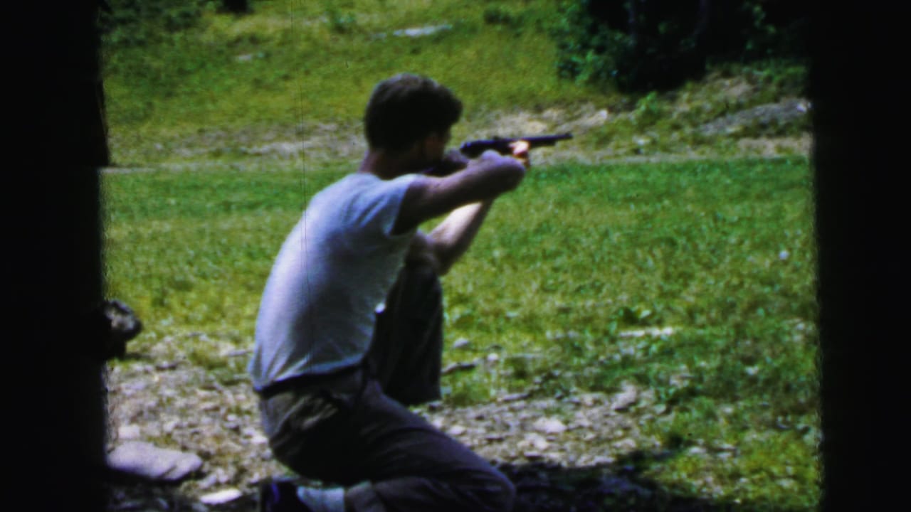 Man in white t-shirt kneels on gravel aiming a handgun toward the right of frame, green grassy hillside behind him.