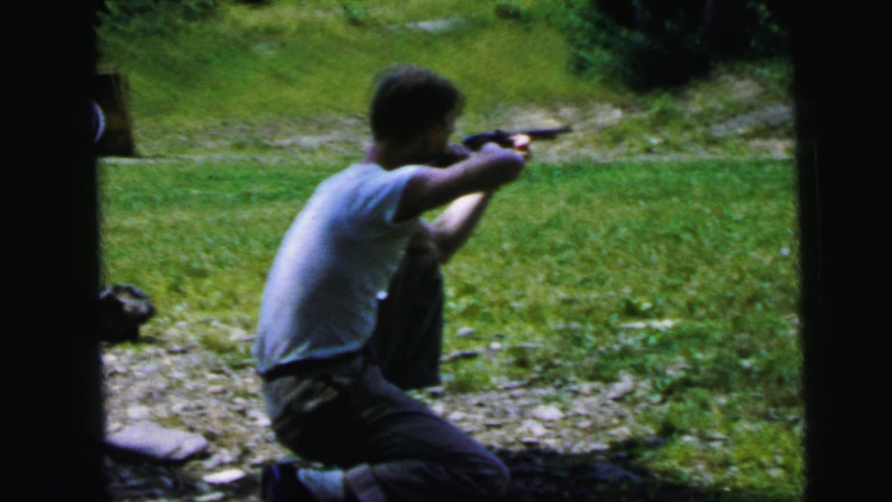 Same man in white t-shirt maintains two-handed aiming stance with handgun, green slope visible behind, slightly shifted position.