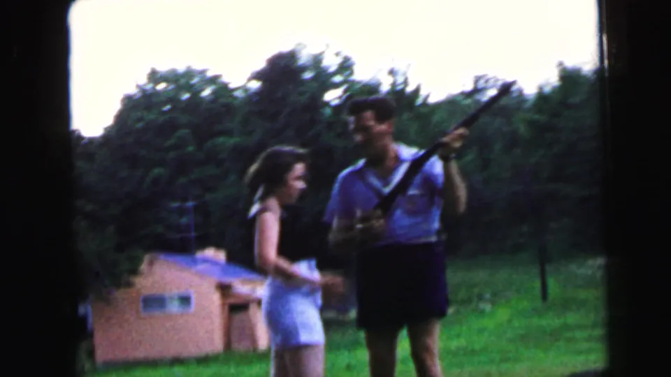 Man holding a rifle upright and woman in white shorts standing on a grassy lawn with a small outbuilding and trees behind them, 8mm home movie circa 1964, Camden, New Jersey.