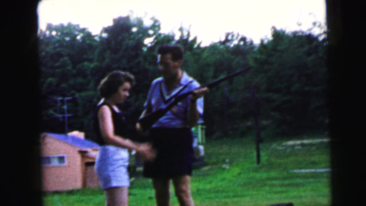 Man and woman standing on a grassy lawn, man holding a long rifle while the woman watches closely.