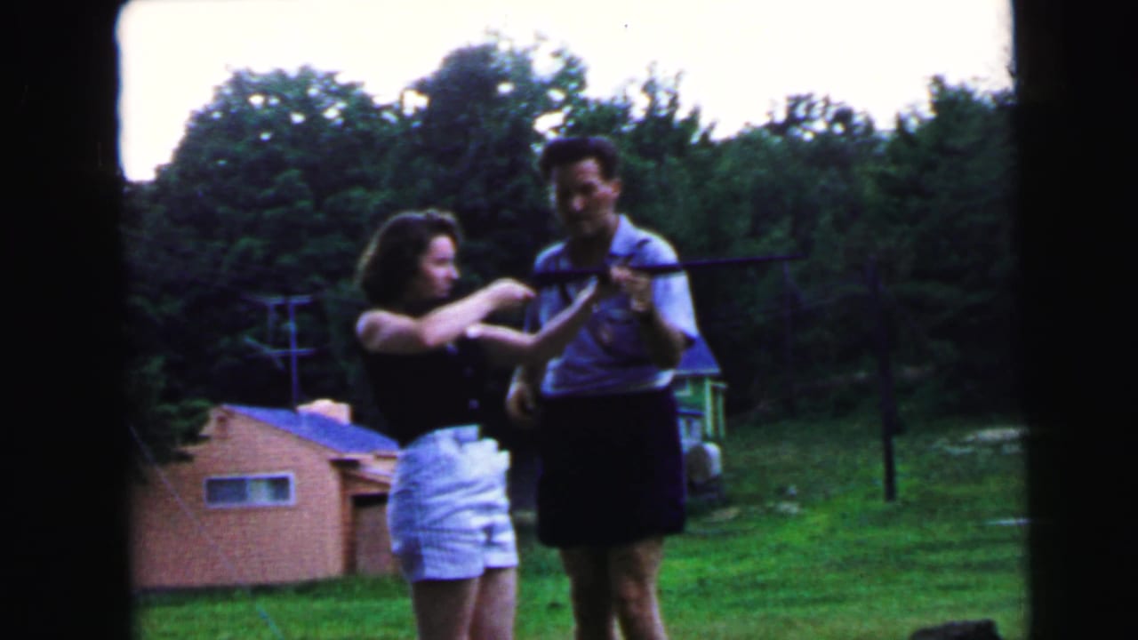 Man in light shirt adjusts or demonstrates rifle hold toward woman in dark sleeveless top and white shorts; suburban backyard with pink outbuilding and utility pole visible, Camden NJ, circa 1964.