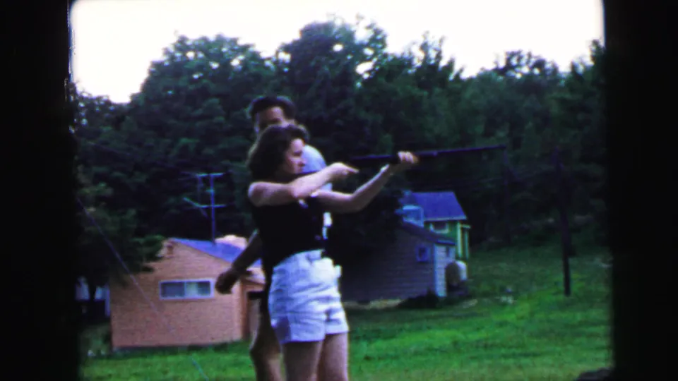 Woman in dark sleeveless top and white shorts raises a long rifle to aim, with man standing close behind coaching her stance; pink and blue-roofed outbuildings visible, suburban yard, 1964.