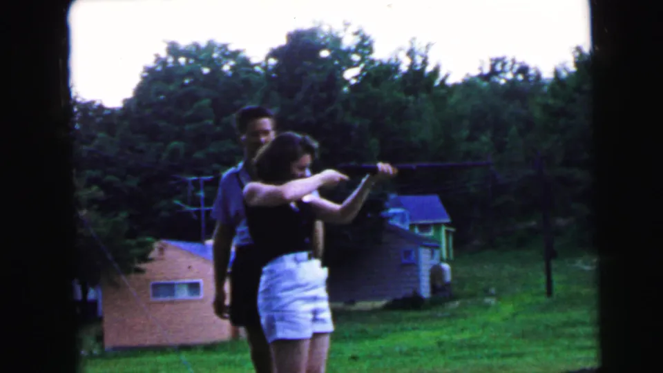 Woman raises rifle to shoulder in shooting position as man coaches from directly behind; pink and blue-roofed outbuildings and dense summer tree line frame the suburban yard, Camden NJ, 1964.