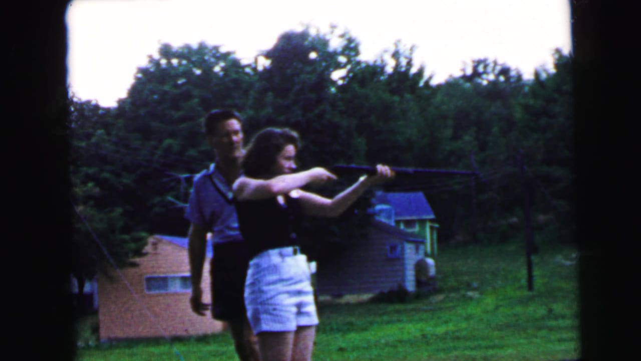 Woman aiming a long rifle in a suburban backyard while a man stands close behind; brick outbuilding and dense trees visible; 8mm home movie, likely Camden NJ, ca. 1964.