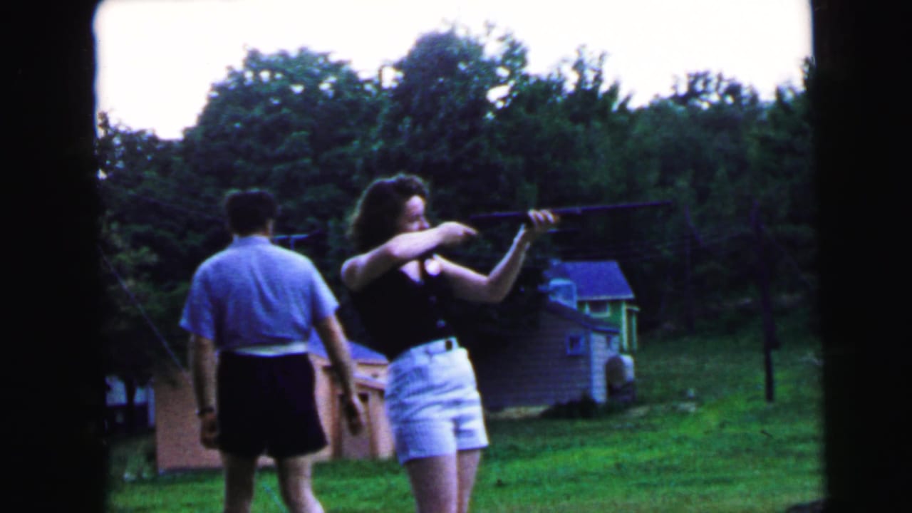 Woman shooting rifle in suburban backyard with man in light blue shirt observing from behind; brick outbuilding and tree line visible; 8mm home movie, likely Camden NJ, ca. 1964.