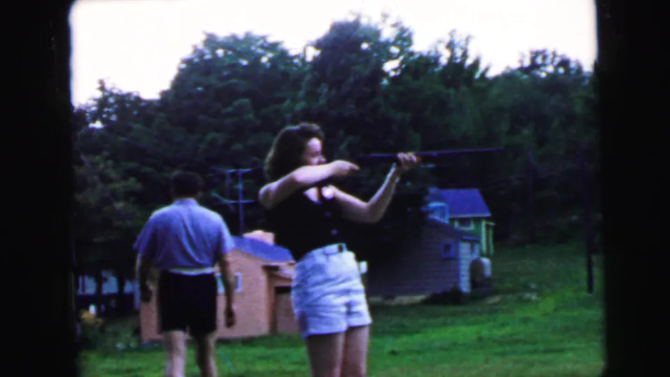 Woman aiming rifle in suburban backyard as man in light blue shirt moves to her left; brick outbuilding with window, small shed, and utility pole visible; 8mm home movie, ca. 1964.