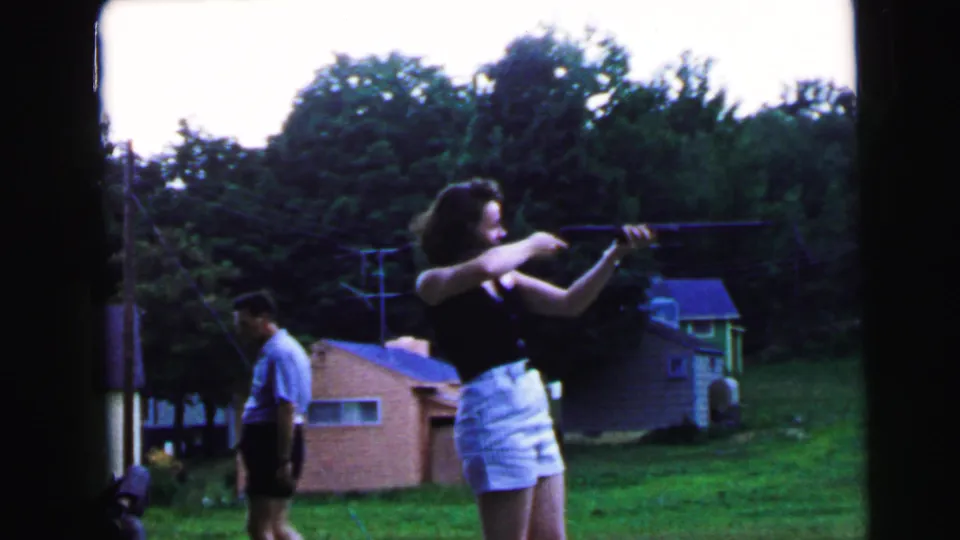 Woman at aim with a long rifle in suburban backyard; man in light blue shirt stepping back to the left; brick outbuilding, utility pole, and dense tree line visible; 8mm home movie, ca. 1964.