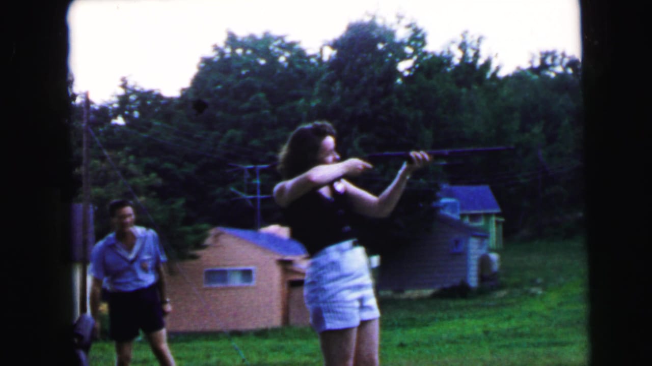 Woman in black top and white shorts aiming a rifle in a grassy backyard; man in light blue shirt watches from behind.