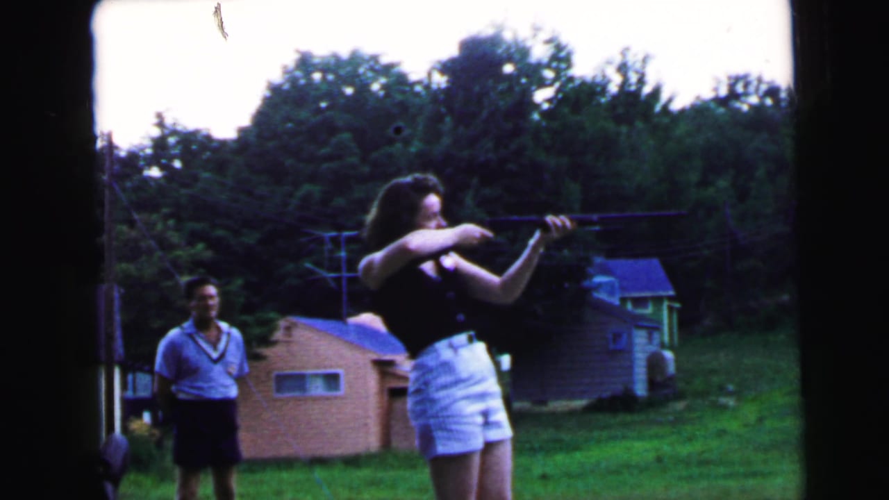 Woman in black top and white shorts continues aiming rifle; man in light blue shirt and dark shorts stands smiling in background.