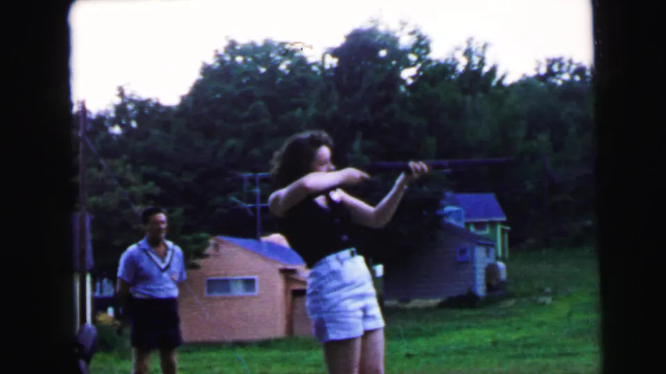 Woman extends rifle in a sustained shooting stance in a 1960s suburban backyard; man watches to her left; outbuildings and tree canopy frame the scene.