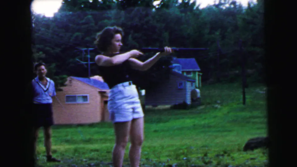 Woman in black sleeveless top and white shorts takes aim with a rifle in a 1960s suburban backyard; male companion smiles behind her; pink brick outbuilding and leafy trees visible.