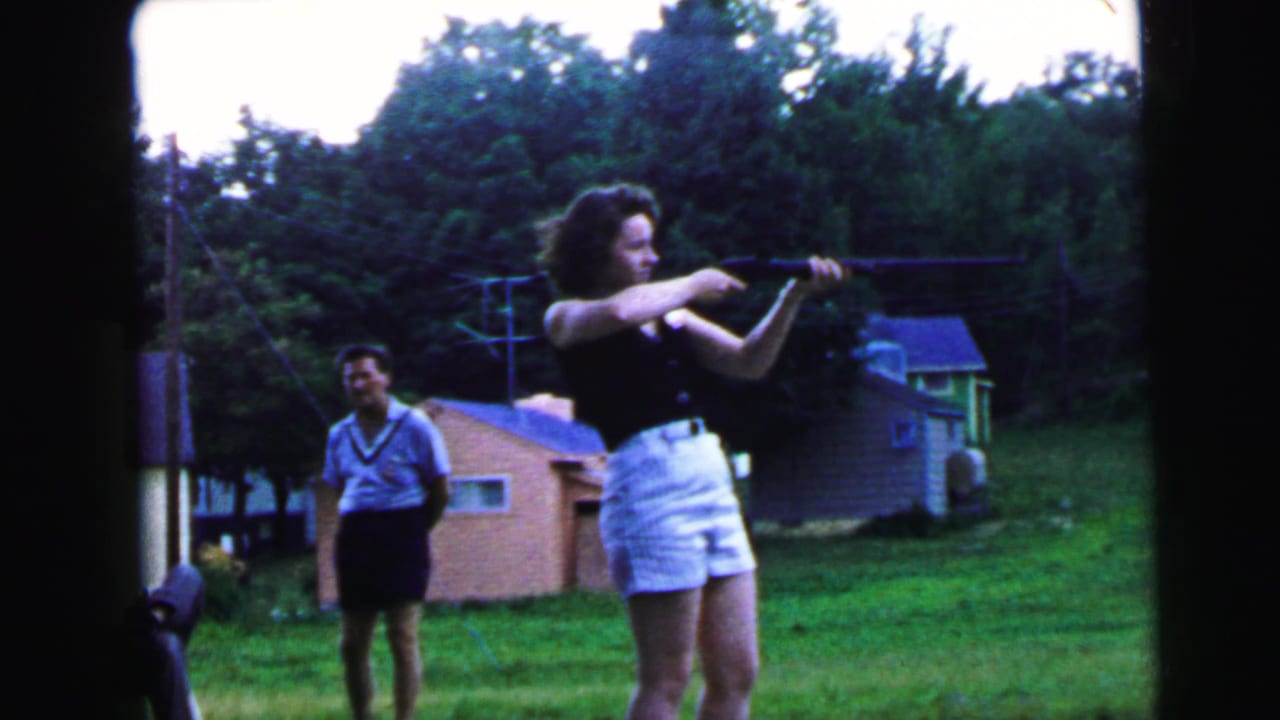 Young woman in black top aims a rifle to the right across a grassy backyard; young man in light blue V-neck shirt watches from behind.