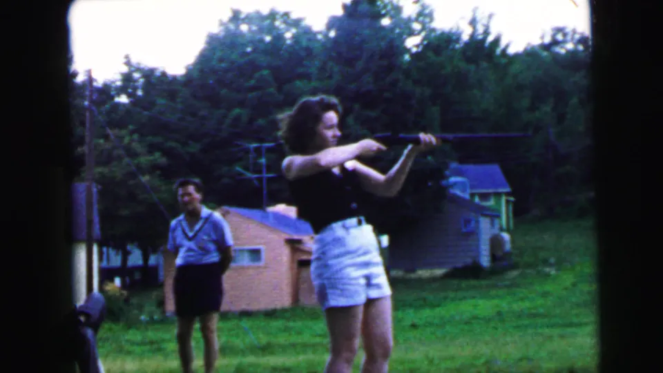 8mm home movie still, c.1964, possibly Camden NJ: young woman aiming a rifle in a suburban backyard while a male companion stands watching.